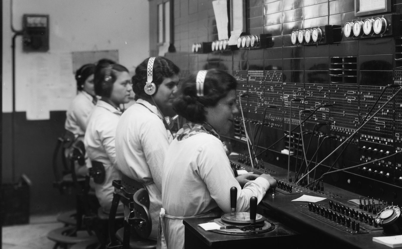 Four women wearing headsets work side by side at a large telephone switchboard.